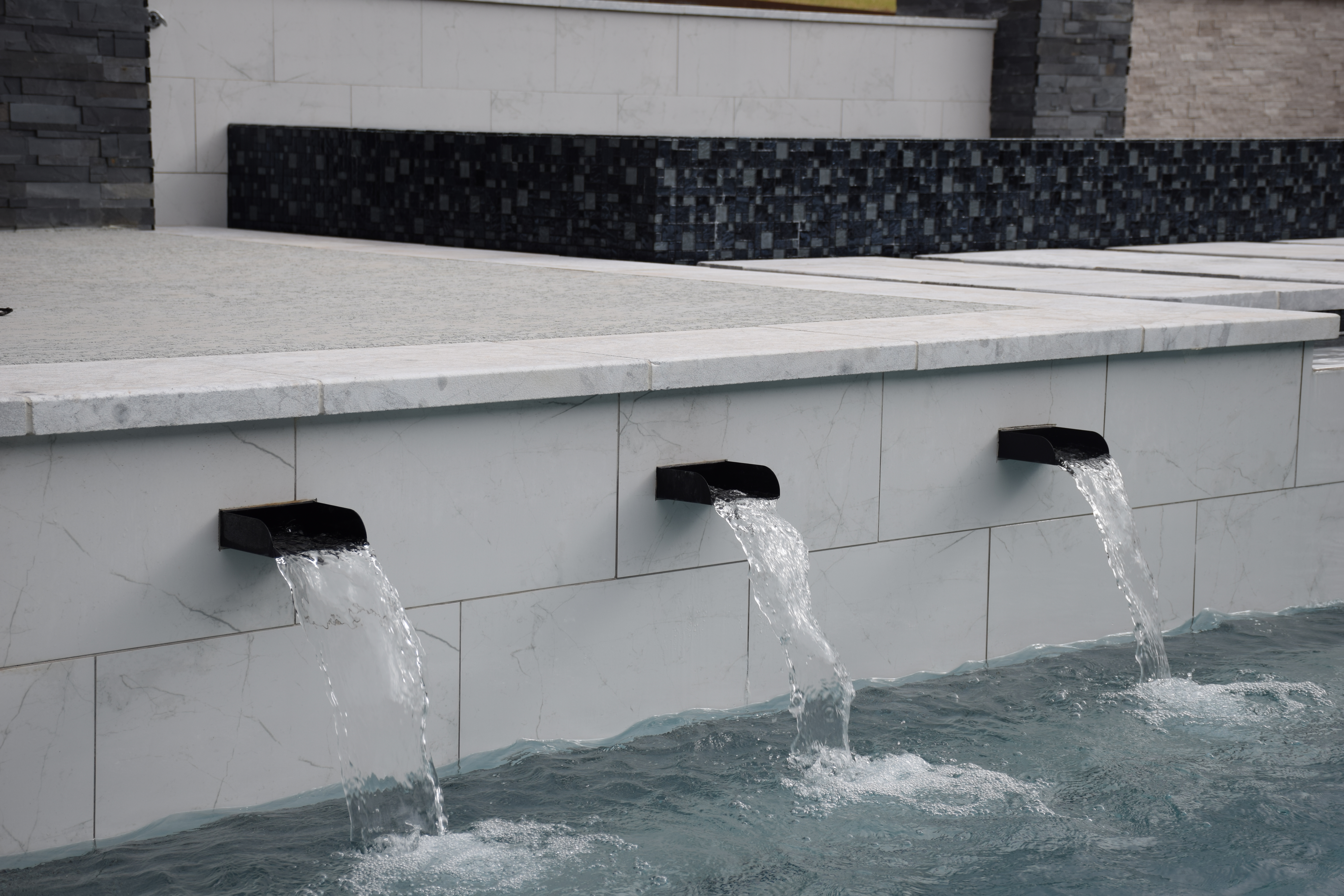 Three black scupper spouts pouring water into a renovated pool with new marble waterline tile