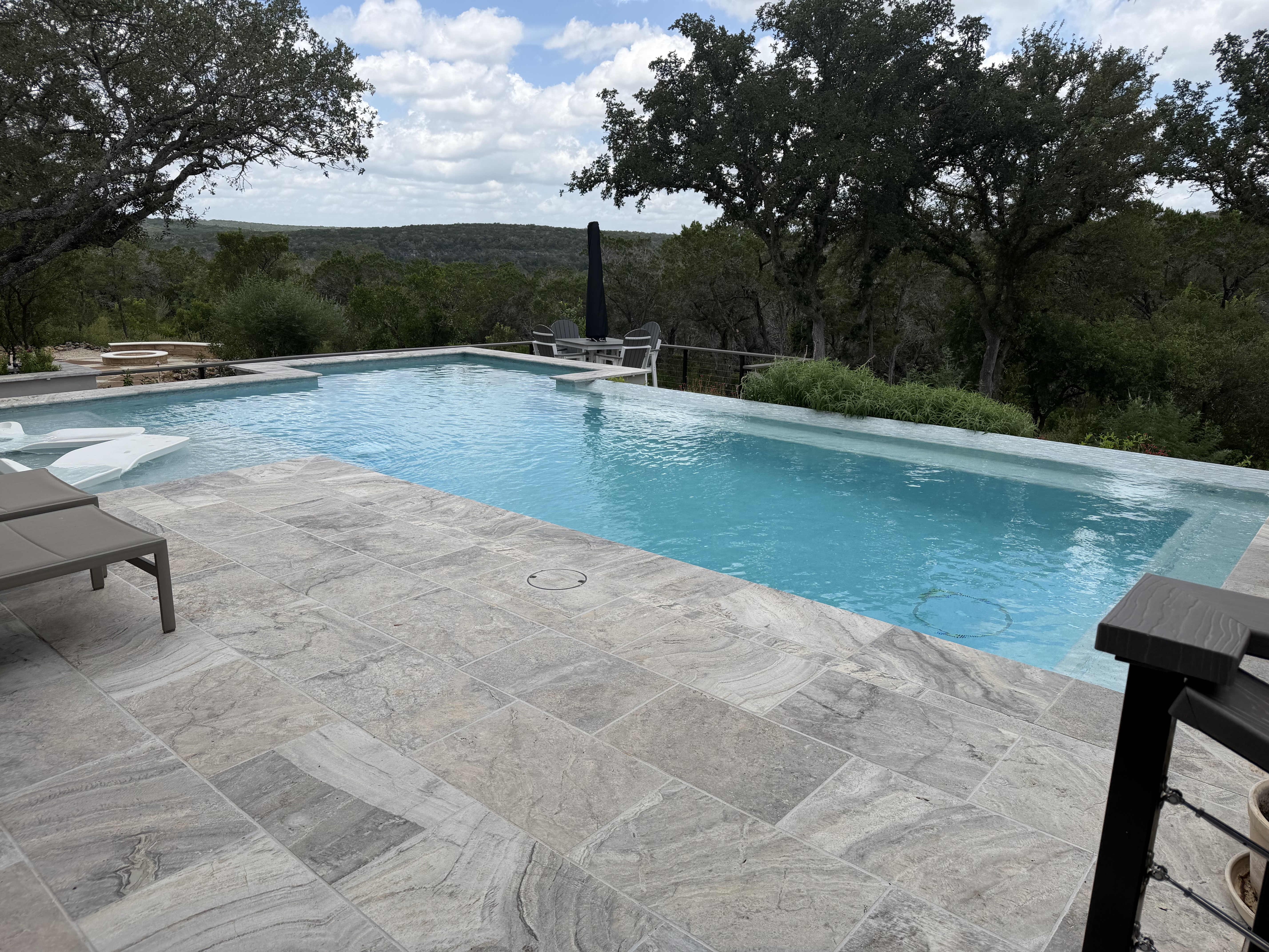 Vanishing edge of the pool disappearing into the Hill Country tree line, with travertine coping and a raised sun shelf