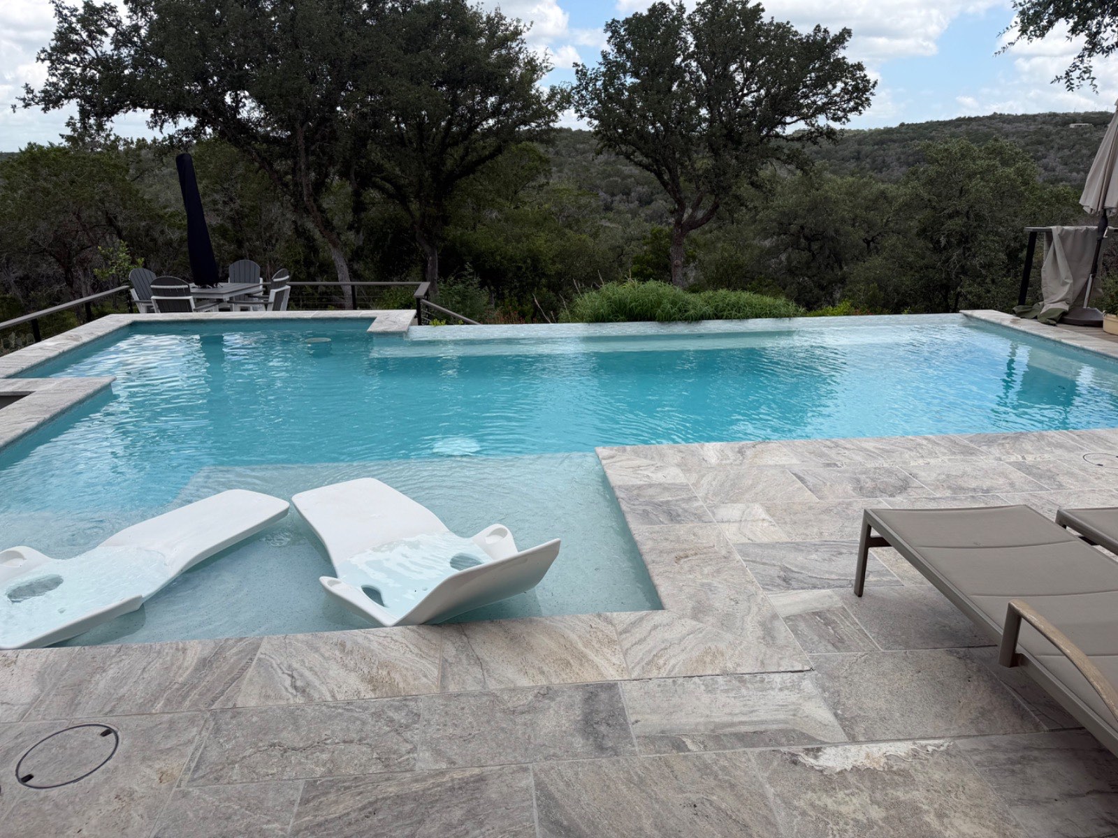 Vanishing-edge custom pool with travertine deck, in-water loungers, and sweeping Texas Hill Country views under a canopy of live oaks