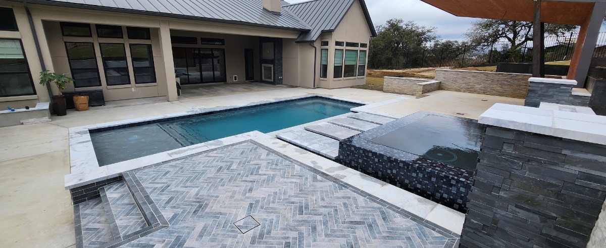 Rectangular custom gunite pool with raised black glass-tile spa, sheer-descent spillway, and herringbone stone decking built by J. Oliver Pools