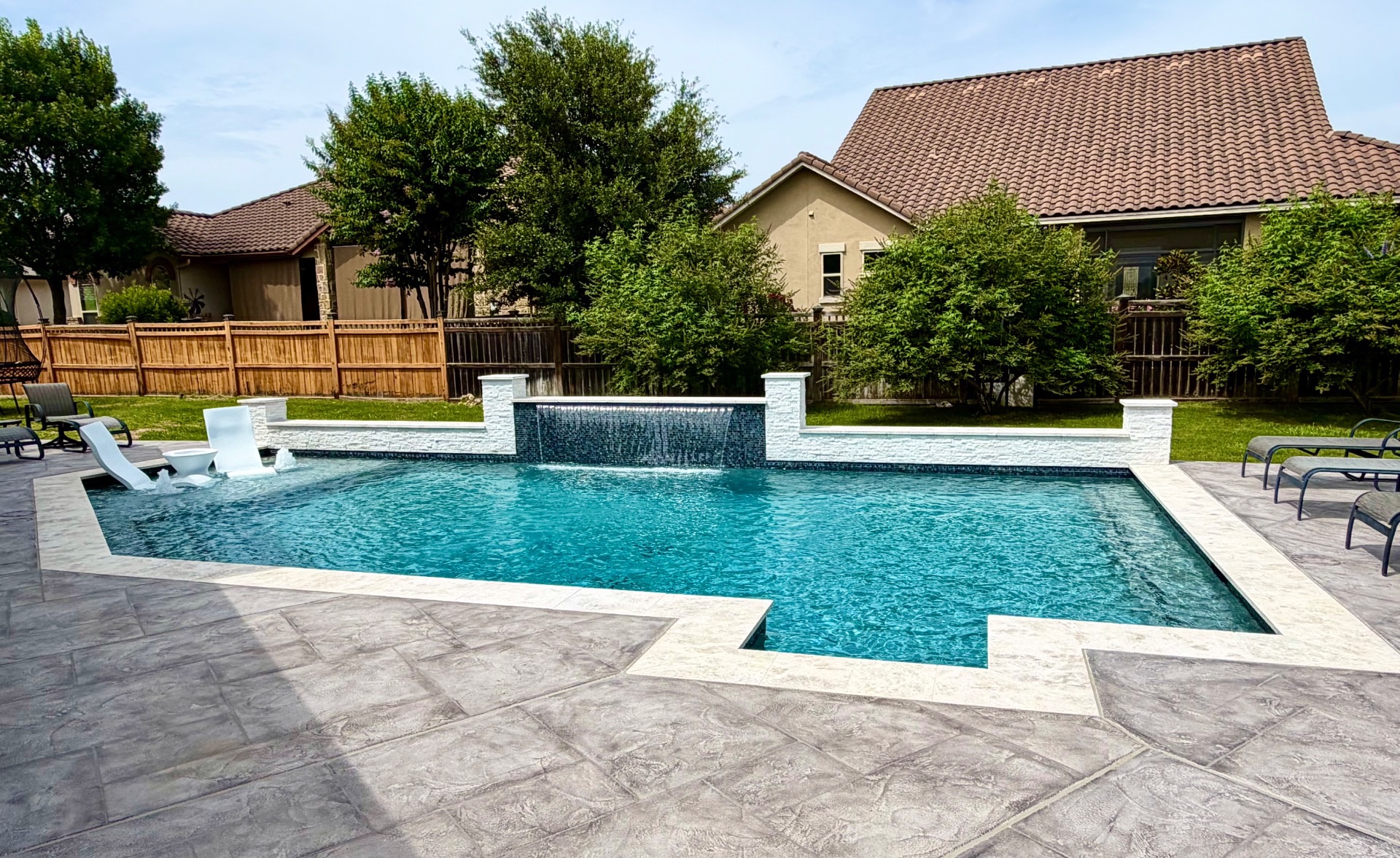 Geometric custom gunite pool with glass-tile sheer-descent water feature, white stacked-stone wall, and travertine deck built by J. Oliver Pools in New Braunfels, Texas