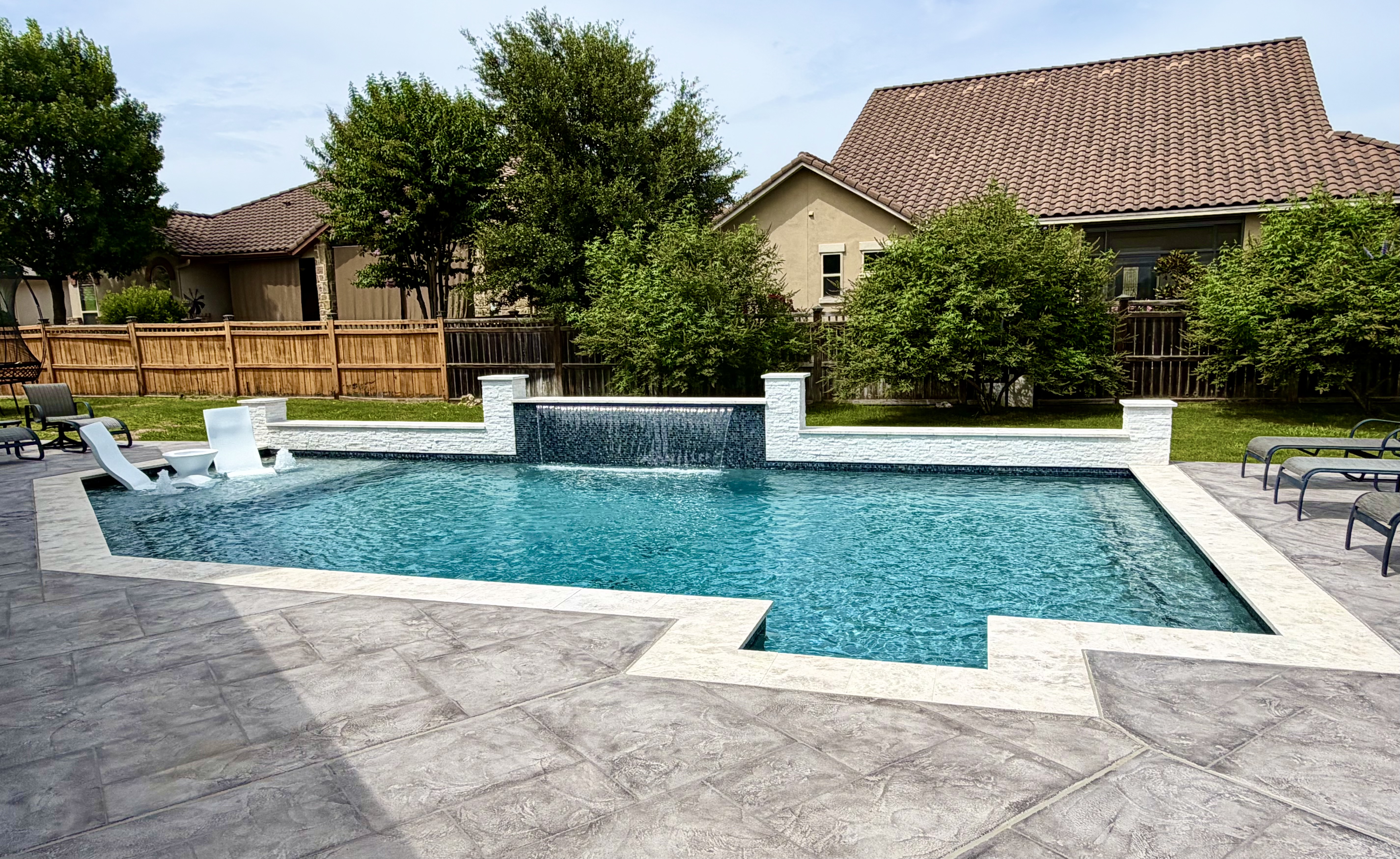 Geometric custom gunite pool with twin glass-tile sheer-descent water features, white stacked-stone wall, tanning ledge, and travertine deck built by J. Oliver Pools in New Braunfels