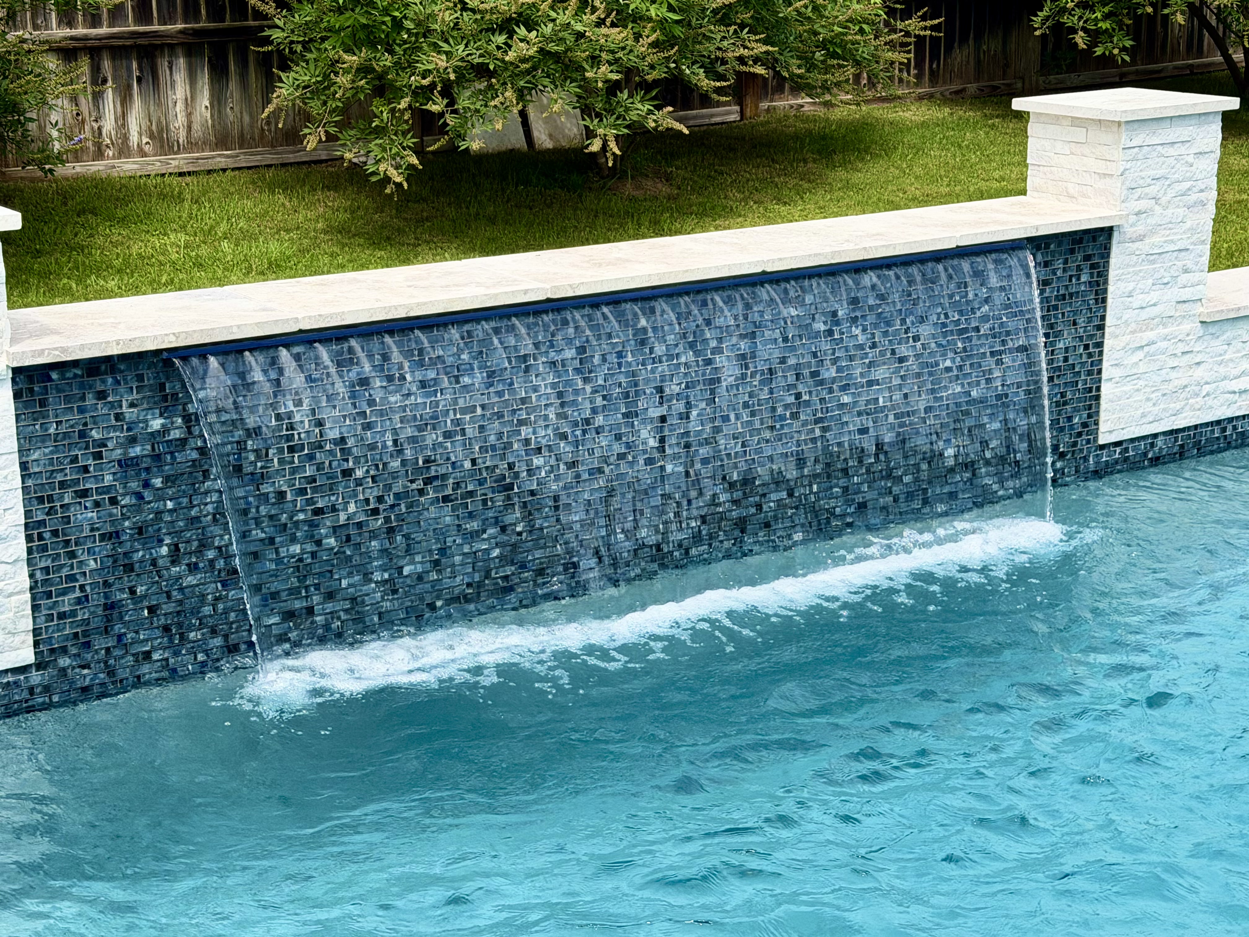 Close-up of a glass-tile sheer-descent waterfall spilling over a deep-blue mosaic wall capped with white limestone in New Braunfels