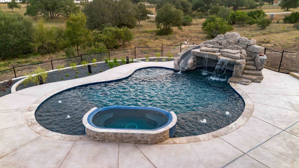 Freeform pool with a rock grotto waterfall, slide, and raised spa overlooking Hill Country ranchland in New Braunfels, Texas