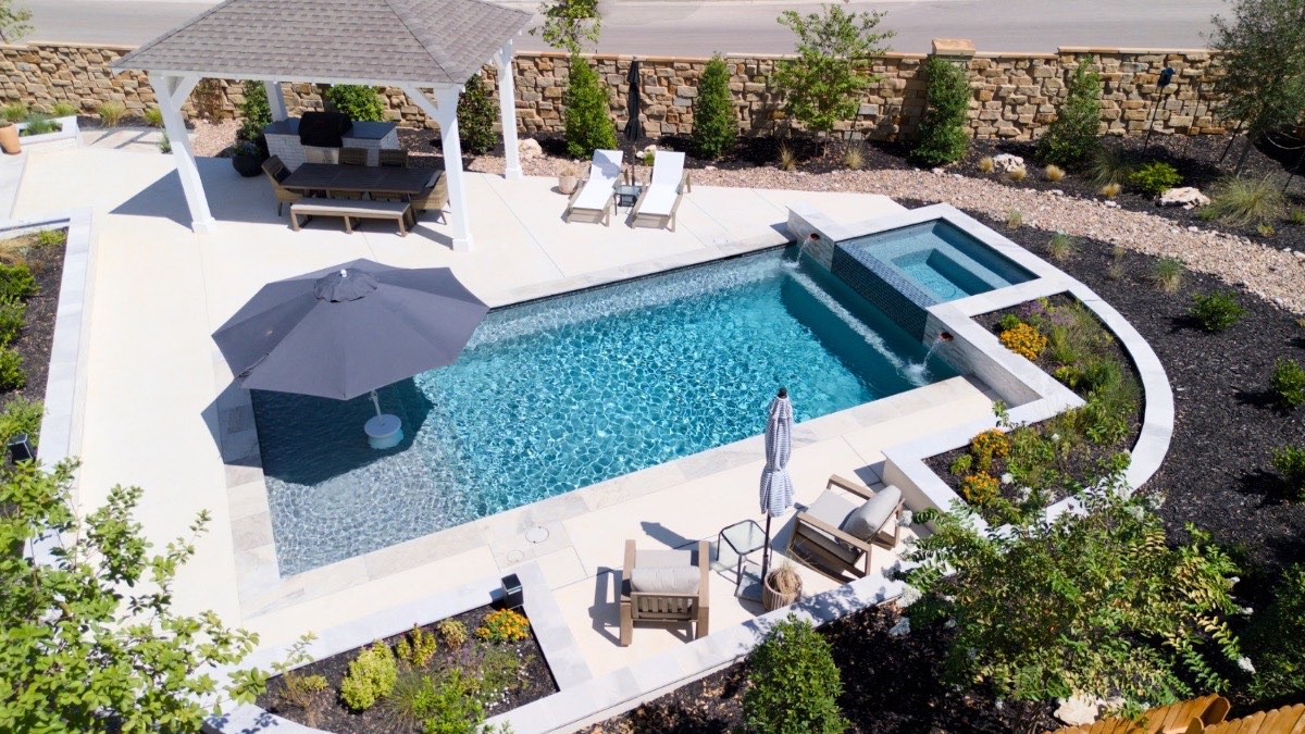 Aerial of a geometric pool and raised spa with pergola, outdoor kitchen, and stone-wall landscaping in New Braunfels, Texas