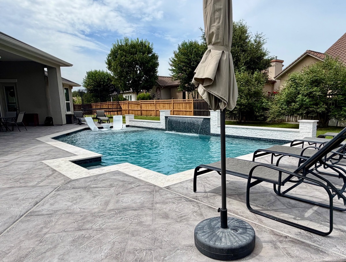 Stamped-concrete patio with loungers, shade umbrella, and travertine pool coping looking across the geometric pool toward the water wall