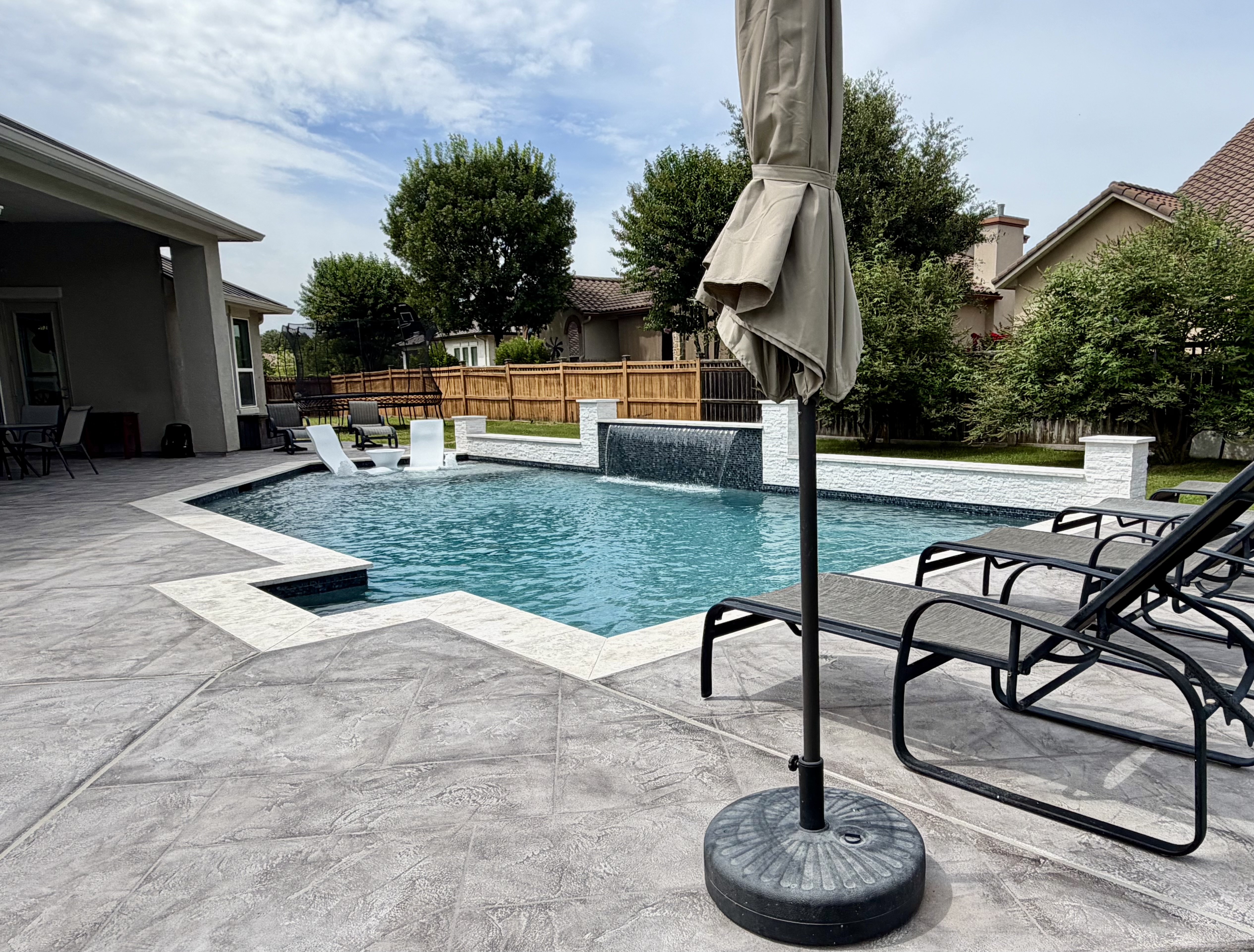 Stamped-concrete patio with loungers, shade umbrella, and travertine pool coping looking across the geometric pool toward the water wall in New Braunfels